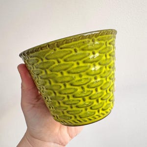 May include: A lime green ceramic planter with a textured basket weave pattern. The rim and base have a darker green trim. The planter is held by a hand against a white background. A decorative item for plants.