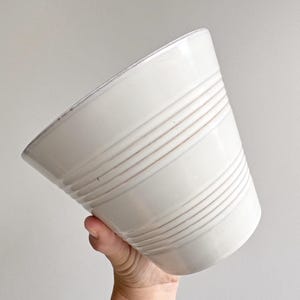 Op de afbeelding: Een witte keramische plantenpot met een taps toelopende, conische vorm en een reeks horizontale, verhoogde ribbels rondom de buitenkant. De pot heeft een gladde, glanzende afwerking en een brede, open bovenkant voor een strak ontwerp.