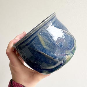 Op de afbeelding: Een handgemaakte keramische kom met een unieke, asymmetrische vorm. De kom heeft een donkerblauwe glazuur met witte plekken en hints van groen en geel. De kom wordt in een hand gehouden, wat de grootte en vorm benadrukt.