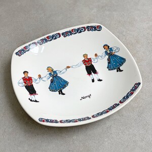 May include: A white ceramic square plate with a blue and white border. The plate features a design of four people in traditional Norwegian folk costumes dancing. The text "Norge" is printed in the center of the plate.