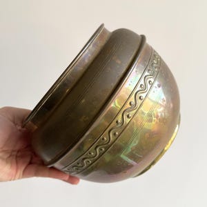 May include: A vintage bronze-toned bowl with a decorative wavy pattern around the middle. The bowl has a rounded shape and a slightly flared rim. The surface has an iridescent sheen, reflecting various colours.