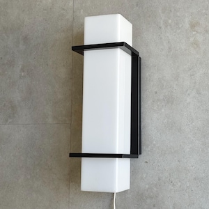 Pode incluir: Arandela de parede moderna com uma luminária branca retangular envolta em uma estrutura de metal preta. A arandela é montada em uma parede cinza e possui um cabo de alimentação branco.