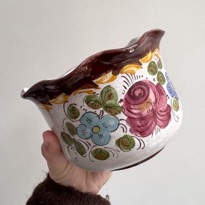Puede incluir: Un cuenco de cerámica blanca con borde marrón oscuro y diseños florales. El cuenco presenta flores pintadas a mano en tonos azules, rosas y verdes. El cuenco está sostenido en una mano.