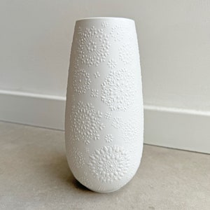 May include: White ceramic vase with a textured design of small, repeating circles and flowers. The vase is tall and slender, with a rounded bottom and a slightly flared opening.
