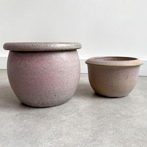 May include: Two ceramic pots, one is a larger, round, light pink pot with a slightly darker rim, and the other is a smaller, round, light brown pot with a slightly darker rim.