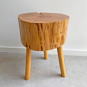 May include: A wooden stool with a circular top and three legs. The stool is made from light brown wood, with a textured surface and a split in the top. The legs are cylindrical. The stool's height is approximately 40 cm.