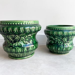 Op de afbeelding: Twee donkergroene keramische plantenpotten met een glanzende afwerking. Elke pot heeft een reliëfpatroon van rozen en bladeren in het midden, met decoratieve kralen- en schulpranden langs de bovenrand en de basis van de vaten.