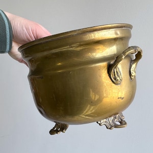 Könnte beinhalten: Ein Vintage-Pflanzgefäß aus Messing mit verzierten Griffen und Füßen. Das Pflanzgefäß hat einen abgerundeten Körper und eine breite Öffnung. Das Messing hat einen warmen, goldenen Farbton mit Gebrauchsspuren, was seinen antiken Charme unterstreicht.