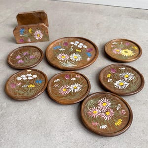 Può includere: Un set di sottobicchieri in legno dipinti a mano e un porta sottobicchieri. I sottobicchieri sono rotondi e ovali, con un motivo floreale di margherite bianche, rosa e gialle. Il porta sottobicchieri ha un design simile. I sottobicchieri misurano circa 10 cm di diametro.