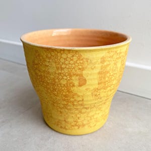 May include: A yellow ceramic planter with a textured, speckled surface. The planter has a wide opening and a slightly tapered shape. The interior is a warm orange color, contrasting with the exterior. Ideal for indoor or outdoor plants.