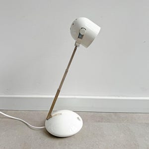 May include: A white desk lamp with a rounded base and a cylindrical head. The lamp has a long, adjustable arm and a white power cord. The lamp's head can be tilted to direct light. The lamp is made of metal and plastic.