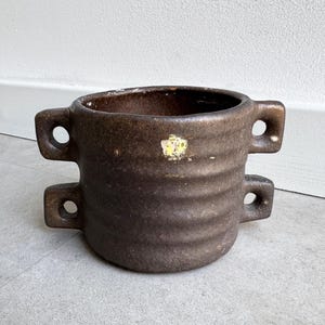 May include: A brown ceramic planter with two sets of handles, each with a circular opening. The planter has a textured, ridged surface and a small, faded yellow sticker. The interior is dark brown.