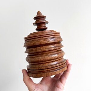 Pu&ograve; includere: Una scatola di legno con un design a strati impilati. La scatola ha una forma rotonda e cilindrica e un coperchio appuntito a forma di guglia.