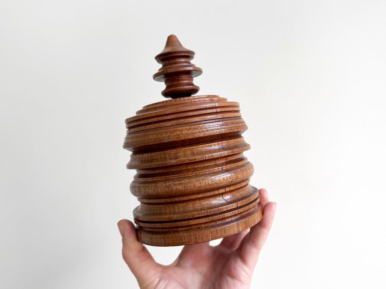 Pu&ograve; includere: Una scatola di legno con un design a strati impilati. La scatola ha una forma rotonda e cilindrica e un coperchio appuntito a forma di guglia.