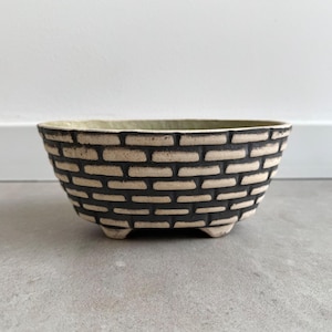 Puede incluir: Una maceta de cerámica blanca con un patrón de ladrillo negro. La maceta tiene cuatro patas pequeñas y es de forma ovalada.