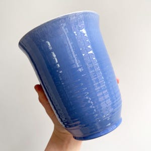 May include: A blue ceramic vase with a textured surface. The vase has a wide opening and a slightly curved shape. The interior rim is white. The vase is held by a person.