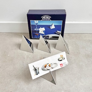 May include: A set of four stainless steel pyramid-shaped place card holders, with a matching box in the background. The box has the word "MENU" on it. A small card with a picture of a champagne bucket and plates is in front of the holders.