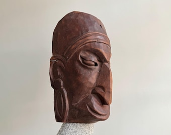 Pode incluir: Uma máscara de madeira castanho-escuro com um rosto esculpido, olhos fechados e um nariz grande e proeminente. A máscara tem um design detalhado e parece ser esculpida à mão.
