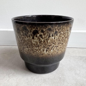 Pode incluir: Um vaso de cerâmica castanho escuro com um design texturizado e salpicado em tons de castanho e bege. O vaso tem um topo largo e ligeiramente alargado e uma base escalonada. O interior é castanho escuro brilhante.