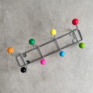 Pode incluir: Um cabideiro moderno com uma estrutura cromada e botões redondos coloridos. Os botões são em várias cores, incluindo vermelho, amarelo, verde, azul, rosa, laranja e preto. Projetado para montagem na parede.