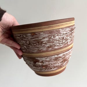 May include: A brown and white ceramic bowl with a textured, layered design. The bowl has a wide opening and a rounded base. The exterior features horizontal bands of brown, beige, and white. The bowl is held by a person.