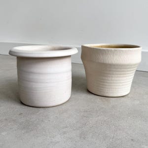 May include: Two off-white ceramic planters. The planter on the left is cylindrical with a wide rim. The planter on the right is a tapered shape with a textured surface. Both planters have a neutral color palette.