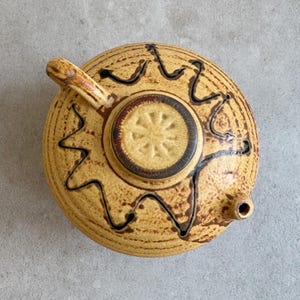 Op de afbeelding: Een bruine en gele theepot van keramiek met een zwart en bruin ontwerp. De theepot heeft een rond lichaam en een tuit aan de zijkant. De deksel heeft een cirkelvormig ontwerp in het midden.