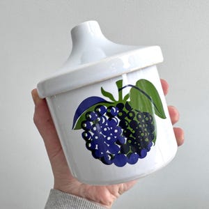 May include: White ceramic jar with a lid, decorated with a painted design of purple grapes and green leaves. The jar is held in a hand, highlighting the fruit motif. A kitchen or home decor item.