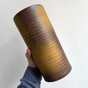 May include: A cylindrical ceramic vase with a textured surface. The vase has a gradient of colors, transitioning from a dark brown at the base to a golden yellow near the top. The vase is held at an angle.