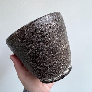 Könnte beinhalten: Eine dunkelbraune Keramikschale mit strukturierter Oberfläche und weißem Sprenkelmuster. Die Schale wird in einer Hand gehalten und hat eine leicht abgewinkelte Form. Der Rand hat eine weiße Umrandung.