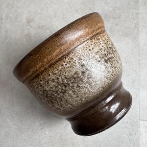 May include: A ceramic bowl with a speckled, textured surface. The bowl has a dark brown base and rim, with a lighter brown and beige speckled body. The bowl has a wide opening and a rounded base.