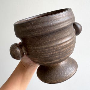 Pode incluir: Um recipiente de cerâmica castanho-escuro com um acabamento mate e texturado. O vaso apresenta uma abertura larga, um corpo arredondado com estrias horizontais, duas pegas laterais esféricas e uma base de pedestal robusta e alargada.
