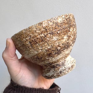 Puede incluir: Un cuenco de cerámica pequeño con base, que presenta una superficie exterior muy texturizada y rugosa. El cuenco tiene una paleta de colores moteados en tonos crema, beige y marrón oscuro, creando un aspecto robusto e irregular.