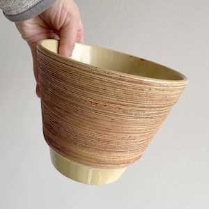 Op de afbeelding: Een beige en bruine keramische kom met een textuur, horizontaal gestreept ontwerp. De kom heeft een brede, schuine opening en een afgeronde basis. De kom wordt door een persoon vastgehouden.