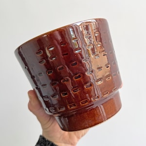 Pode incluir: Um vaso de cerâmica marrom com uma superfície texturizada. O vaso tem uma borda ligeiramente alargada e um fundo arredondado.