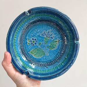 May include: A round, blue ceramic ashtray with a floral design. The ashtray has a textured surface and a central flower motif in shades of blue and green. It has three notches for resting cigarettes.