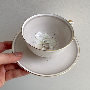 May include: A delicate teacup and saucer set. The cup and saucer are a soft beige with gold trim. The cup features a floral design inside, and the saucer has small gray dots. A classic and elegant tea set.