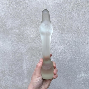 Könnte beinhalten: Eine dekorative Flasche aus mattiertem Glas mit rundem Boden und langem Hals. Die Flasche wird in einer Hand vor einem grau strukturierten Hintergrund gehalten. Das Glas hat ein weiches, durchscheinendes Aussehen.