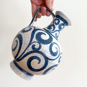 May include: A ceramic pitcher with a round body and a handle. The pitcher is decorated with a blue and white swirl pattern. The pitcher has a spout and a base. The pitcher is being held by a hand.