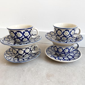 May include: Four ceramic teacups and four matching saucers arranged in two stacks. Each piece features a cream-colored base with a repeating pattern of dark blue circular outlines and small grey-green centers. The cups have curved handles.