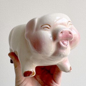 Puede incluir: Figura de cerdo de cerámica vintage, principalmente blanca con detalles rosas en el hocico, las orejas y las mejillas. El cerdo tiene una expresión sonriente y detalles marrones en las pezuñas. Un objeto decorativo encantador.