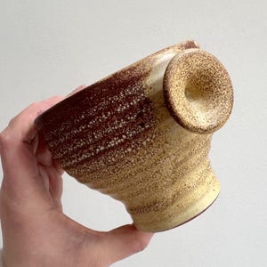 May include: A handcrafted ceramic bowl with a distinctive design. The bowl has a speckled, textured surface with a gradient from brown to beige. It features a unique handle and a flared rim, ideal for serving or display.