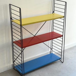 Könnte beinhalten: Ein dreistufiges Regal mit einem schwarzen Metallrahmen. Die Regale sind in Gelb, Rot und Blau lackiert. Das Regal hat ein minimalistisches Design und eignet sich zum Ausstellen von Gegenständen in einem Haus oder Büro. Die Regale sind ca. 70 cm lang.