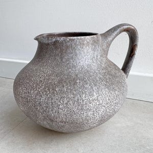 Può includere: Una brocca in ceramica con un corpo rotondo e bombato e un manico curvo. L'esterno presenta una finitura maculata nei toni del marrone e del bianco, creando un aspetto strutturato. Il bordo include un piccolo beccuccio per versare.