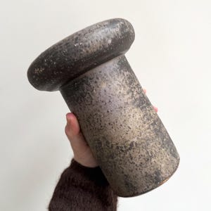 Könnte beinhalten: Eine zylindrische Keramikvase mit breitem, abgerundetem Oberteil und strukturierter Oberfläche. Die Vase hat eine dunkle, gesprenkelte Oberfläche mit Grau- und Brauntönen. Die Vase wird vor einem weißen Hintergrund gehalten.