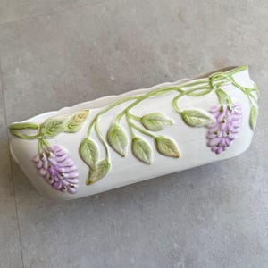 以下が含まれることがあります： 長方形のオフホワイトのセラミックプランター。緑の葉と紫の藤の花のレリーフが施されています。縁は緑色のトリムでアクセントになっています。