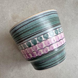 Pode incluir: Um vaso de cerâmica com um design turquesa e roxo. O vaso apresenta listras pretas horizontais e uma faixa de formas de gota verdes e roxas alternadas. A borda é de cor castanho claro.