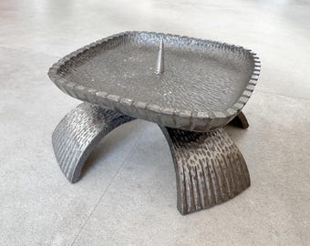 May include: A silver metal candle holder with a square base and three arched legs. The holder has a textured surface and a single spike in the center.