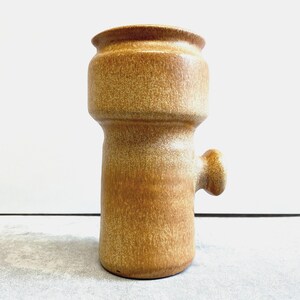 May include: A vintage, tan-colored ceramic vase with a unique, cylindrical shape. The vase features a rounded top, a wider midsection, and a small, rounded handle on the side. The surface has a speckled texture, adding to its rustic charm.