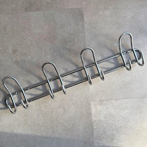 May include: A wall-mounted coat rack made of polished chrome metal, featuring four double-pronged hooks arranged in a horizontal row. Each hook has a rounded, U-shaped design with a smaller secondary hook extending from the base of the main arm.